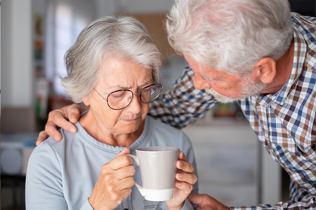 Un vieil homme prend soin d'une vieille dame. Cette dernière tient un tasse dans ses mains.