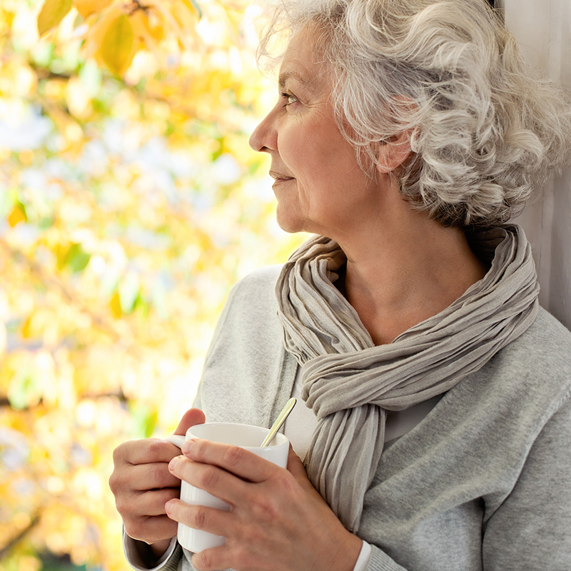 Une femme âgée est appuyée sur un mur. Elle tient une tasse de café dans ses mains et regarde au loin.