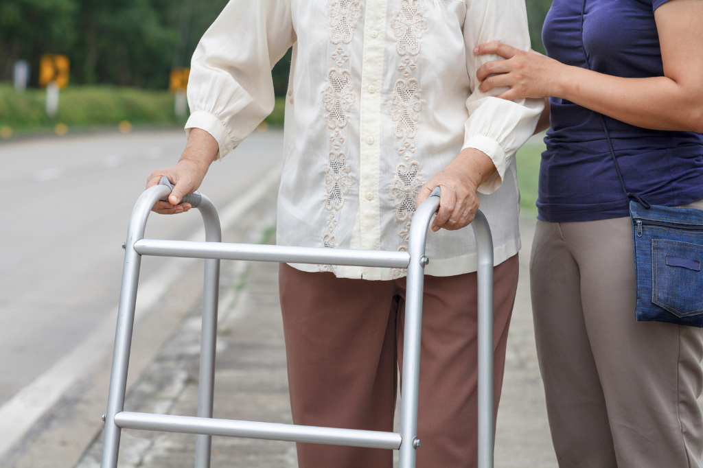 Une femme accompagnée une aînée qui marche avec un déambulateur sur le trottoir.